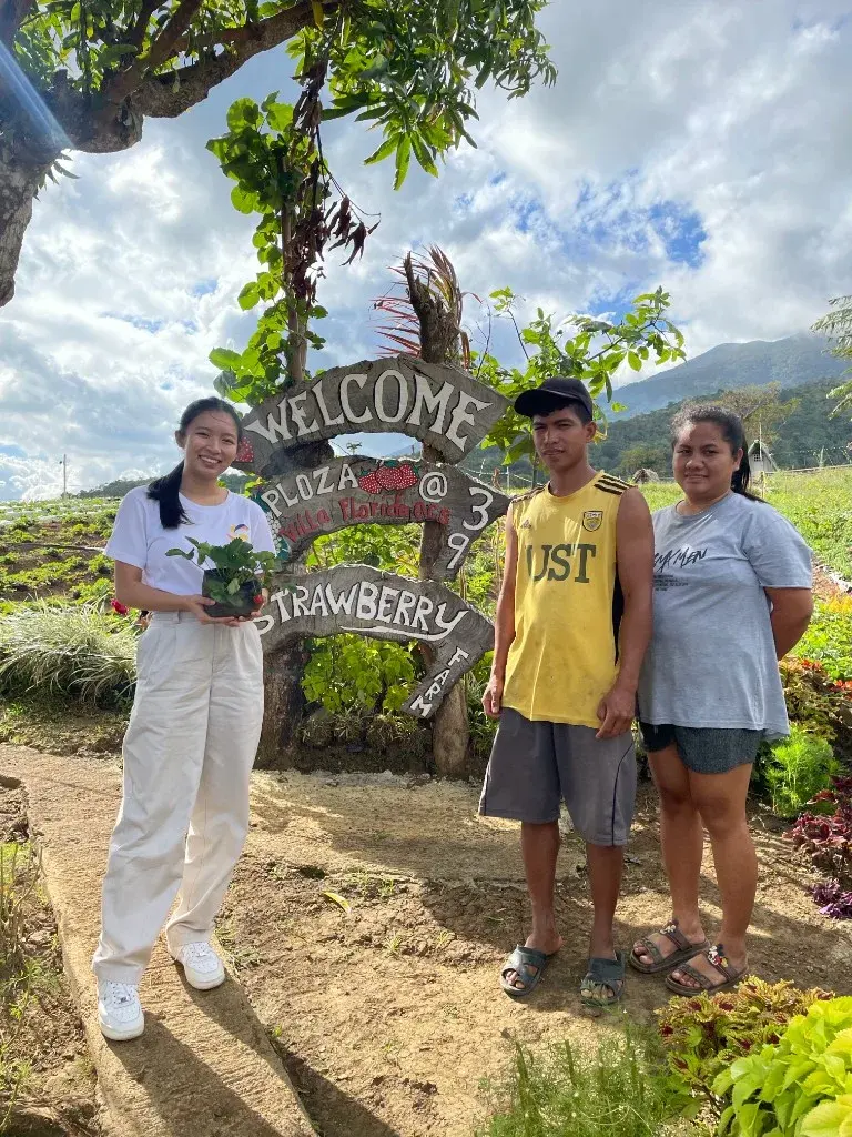Three hosts beside a wooden welcome sign at a strawberry farm, with fields and mountains behind them